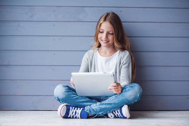 Αρκετά εφηβικό κορίτσι είναι χρησιμοποιώντας ένα ψηφιακό tablet και χαμογελώντας ενώ κάθεται στο πάτωμα - Φωτογραφία, εικόνα