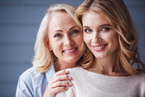 Beautiful senior mom and her adult daughter are hugging, looking at camera and smiling - Photo, Image