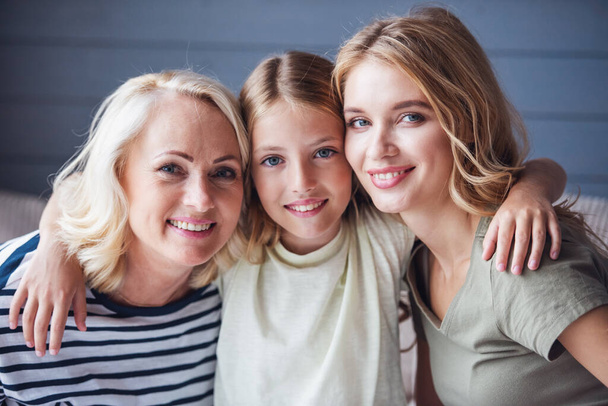 Beautiful women generation: granny, mom and daughter are hugging, looking at camera and smiling while sitting on couch at home - Photo, Image