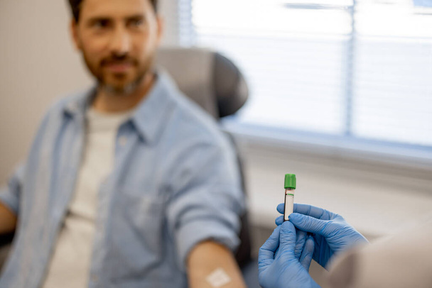 Laboratoriumassistent houdt buis met bloed voor analyse, gelukkige mannelijke patiënt op de achtergrond, focus op een buis - Foto, afbeelding