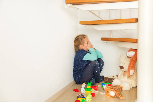 Девушка сидит в углу под лестницей и в страхе закрывает лицо руками, плачущим одиноким ребенком и детскими игрушками. Детские страхи, психологические проблемы детей. - Фото, изображение