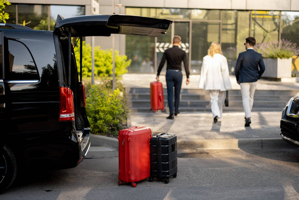 Řidič nebo concierge pomáhá obchodnímu páru nést své kufry do kanceláře nebo hotelu z minivanu taxi. Koncepce služebních cest a dopravní služby - Fotografie, Obrázek