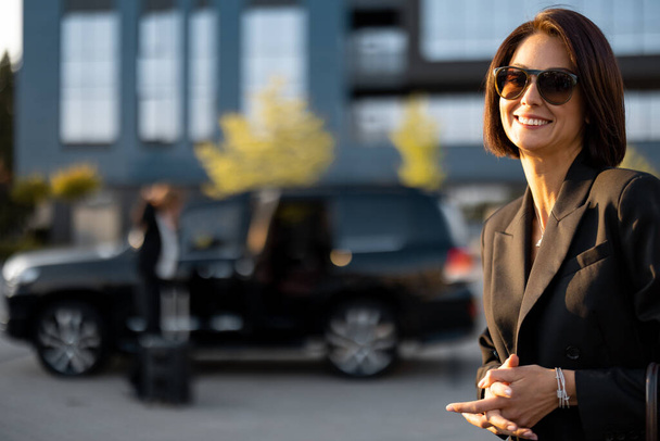 Muotokuva hymyilevä liiketoiminnan nainen musta muodollinen kulumista ja aurinkolasit ulkona, auton ja toimistorakennus taustalla - Valokuva, kuva