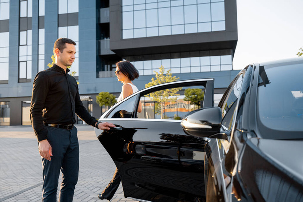 Chauffeur ajuda uma mulher de negócios elegante a entrar no carro, abrindo a porta de um táxi premium perto de um prédio de escritórios ou hotel. Conceito de motorista pessoal ou viagens de negócios - Foto, Imagem
