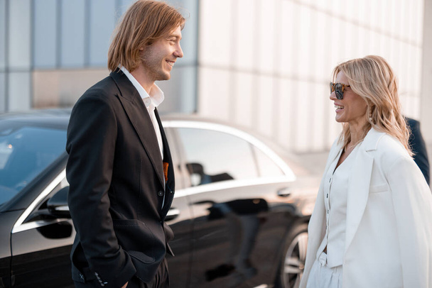 Eleganccy biznesmeni rozmawiają w pobliżu luksusowego samochodu na zewnątrz. Partnerzy biznesowi rozmawiają po wieczornej imprezie na parkingu - Zdjęcie, obraz