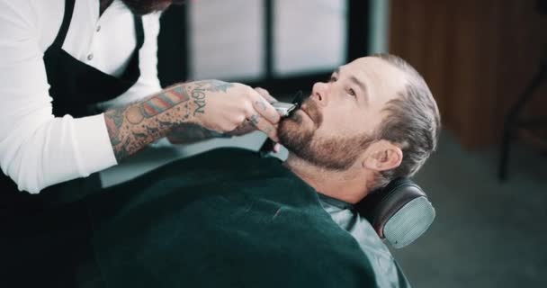 Beard, shave or salon with a barber and man in a seat as a customer for luxury or professional service. Industry, grooming or hairdresser and a person shaving the face of a client with a machine. - Footage, Video