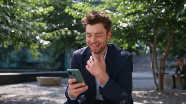 Hombre de negocios chateando teléfono inteligente sentado calle verde de cerca. Sonriente gerente guapo viendo las redes sociales en el teléfono celular al aire libre. Trabajador de oficina feliz relajarse con el teléfono móvil en el descanso del trabajo. - Imágenes, Vídeo