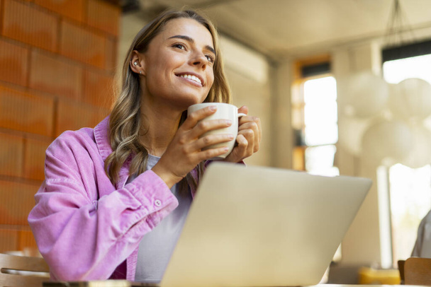 Ritratto sorridente bella donna che utilizza computer portatile bere caffè seduto in ufficio moderno. Successo freelancer pianificazione startup progetto guardando altrove sul posto di lavoro o caffè. Concetto pausa caffè  - Foto, immagini
