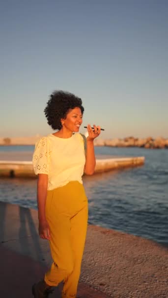 Video van een Afrikaanse vrouw die glimlacht tijdens het opnemen van een voicemail met de mobiele telefoon naast de zee tijdens zonsondergang - Video