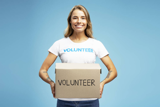 Hymyilevä vapaaehtoinen nainen, jolla on pahvilaatikko, jossa lahjoitukset katselevat sinisellä taustalla olevaa kameraa. Hyväntekeväisyyskeskuksen käsite  - Valokuva, kuva