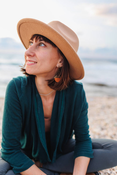 Gyönyörű természetes szépség nő mosolyog és nevet, pózol a kamera a strandon. Portré egy fiatal mosolygós lányról kalapban, egy boldog utazóról, aki a parton ül.. - Fotó, kép