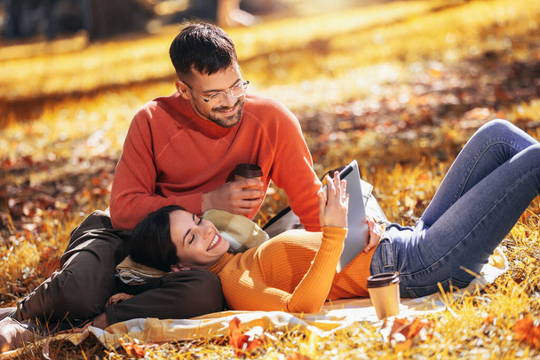 Młody mężczyzna i kobieta siedzą na kocu w parku jesienią za pomocą tabletu cyfrowego. - Zdjęcie, obraz