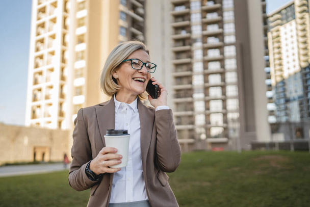 Jedna dojrzała kobieta na zewnątrz przed budynkami rozmawiająca przez telefon komórkowy i trzymająca gotową kawę, nowoczesny biznes i koncepcję stylu życia - Zdjęcie, obraz