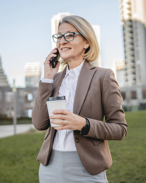 Una mujer madura al aire libre frente a edificios hablando por teléfono móvil y sosteniendo un café para llevar, negocio moderno y concepto de estilo de vida - Foto, Imagen