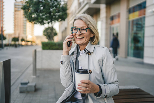 Una mujer madura al aire libre frente a edificios hablando por teléfono móvil y sosteniendo un café para llevar, negocio moderno y concepto de estilo de vida - Foto, imagen