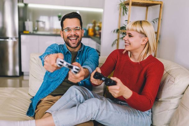 リビングルームでジョイスティックでビデオゲームをプレイするボーイフレンドとガールフレンド。愛するカップルは自宅でビデオゲームをプレイしています - 写真・画像