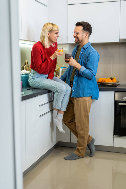 Молодой человек и женщина стоят на кухне и разговаривают. Счастливая жена и муж с чашками кофе и соком стоя на кухне. - Фото, изображение