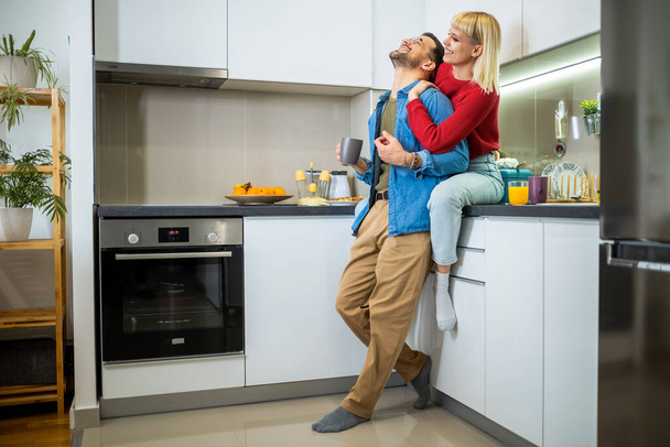 Młody mężczyzna i kobieta stojący w kuchni i mówiący. Szczęśliwa żona i mąż z filiżanek kawy i soku stojących w kuchni. - Zdjęcie, obraz