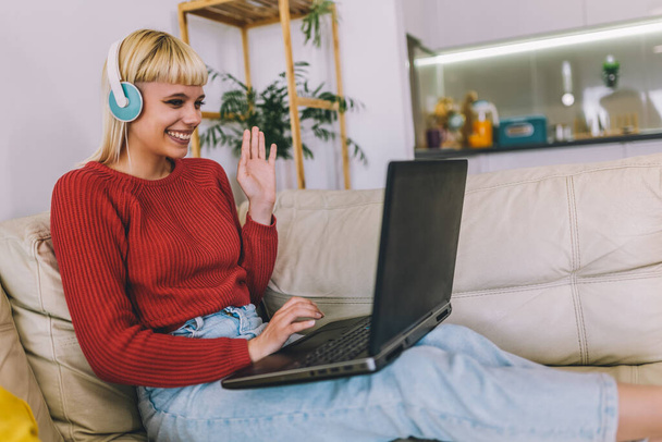 Mujer sonriente usando un portátil para una videollamada mientras está sentada en el sofá en casa. Mujer usando el ordenador portátil y tener una reunión en línea mientras trabaja desde casa - Foto, Imagen