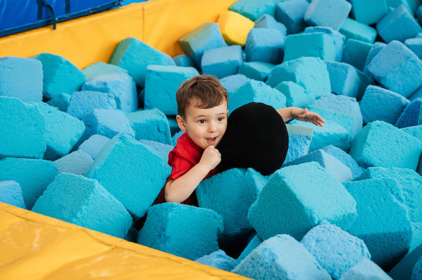 A boy is playing in a game entertainment nursery. He plays with soft large cubes. Active pastime in the amusement park. Trampolines and a dry pool - Фото, изображение