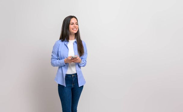 Confiant jeune femme entrepreneure messagerie sur téléphone portable et regardant loin sur fond blanc - Photo, image