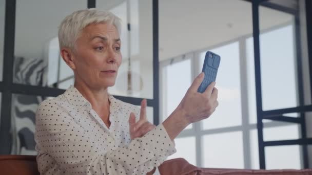 Engaged senior woman with short white hair making a video call on her smartphone, sitting comfortably on a leather couch in a well-lit modern living room - Felvétel, videó