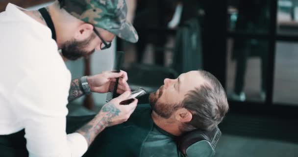 Beard, shave or styling with a barber and man in a seat as a customer for luxury or professional service. Facial grooming, salon or hairdresser and person shaving the face of a client with a machine. - Footage, Video