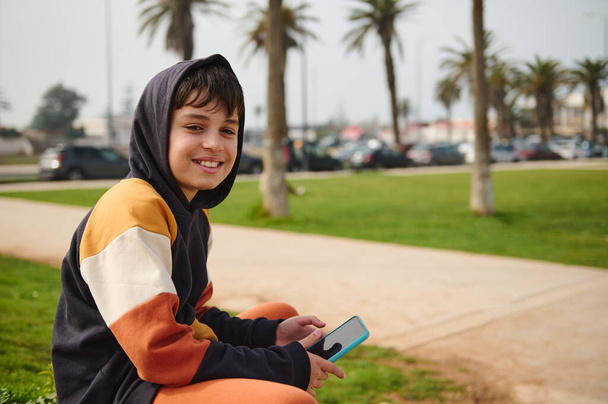 Caucasian happy hipster preadolescent boy smiles looking at camera, holding smart mobile phone while enjoys his weekend outdoor. Online communication, People and internet technologies - Photo, Image