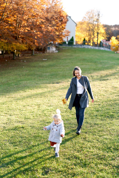 Mama mit einem Strauß gelber Blätter folgt einem kleinen Mädchen, das durch eine grüne Wiese läuft. Hochwertiges Foto - Foto, Bild