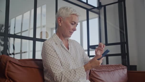 Focused senior woman using a smartphone in a modern living room setting - Felvétel, videó