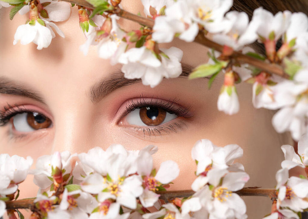 美しいヌードメイク,太い眉毛を持つ少女. 美しいヘーゼル目が近づいています. 近くには美しい咲く春の桜が咲いています. カメラを見ている. プロフェッショナルメイクと化粧品 - 写真・画像