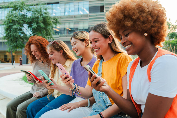 Група усміхнених студентів багаторасового коледжу жінок, які дивляться додаток соціальних мереж або спілкуються зі своїми мобільними телефонами, сидячи за межами університетського містечка. Дівчата-підлітки використовують телефони для розміщення фотографій - Фото, зображення