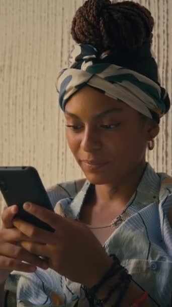 Primer plano vertical de la joven dama afroamericana alegre en durag mirando el teléfono y sonriendo mientras charla en el interior - Metraje, vídeo