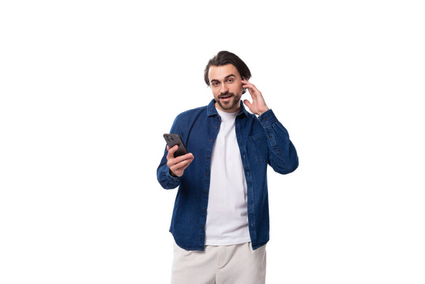 jeune homme brune bien entretenu dans une chemise bleue décontractée tient un téléphone portable dans ses mains. - Photo, image