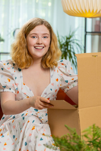 Ευτυχισμένη γυναίκα ξεπακετάρει πακέτο παράδοσης κάθονται στο σπίτι διαμέρισμα στον καναπέ. Χαμογελώντας ικανοποιημένη κοπέλα αγοραστής online κατάστημα πελάτη άνοιγμα χαρτονένιο κουτί παραλαβή δώρο αγοράς από γρήγορη ταχυδρομική αποστολή. Κάθετη - Φωτογραφία, εικόνα