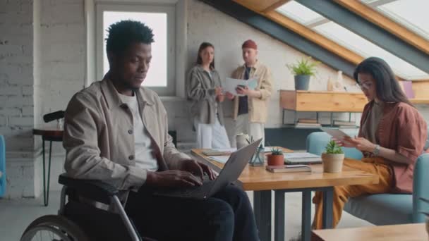 Medium shot van diverse coworking office klanten druk op het werk - zwarte man in rolstoel typen op laptop, Aziatische vrouw met behulp van tablet, en Kaukasische meisje en man bespreken rapport of presentatie - Video