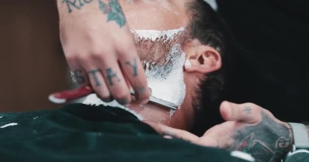 Face, beard or shaving with a barber and man in a seat as a customer for luxury or professional service. Hands, salon or foam and a person grooming the face of a client with a minora blade closeup. - Footage, Video