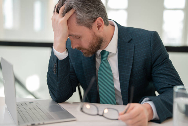 Az Y-generációs üzletembernek fáj a feje. Egy öltönyös üzletember laptopon dolgozik az irodában, szemüveget vesz le migréntől vagy fejfájástól szenvedve. Fáradt, kimerült férfi szenved fejfájás - Fotó, kép