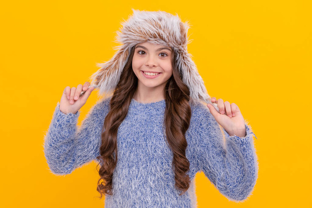 winter fashion. happy kid with curly hair in earflap hat. female fashion model. teenager girl in knitwear on yellow background. portrait of child wearing warm clothes. express positive emotion. - Photo, Image