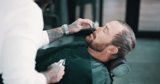 Beard, shave or hairdresser with a barber and man in a seat as a customer for luxury or professional service. Industry, salon or grooming and a person shaving the face of a client with a machine. - Footage, Video