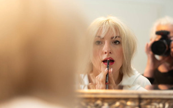 Ξανθιά γυναίκα στέκεται στον καθρέφτη του μπάνιου και εφαρμόζει make-up, ζωγραφίζει το περίγραμμα των χειλιών της με ένα lip liner ενώ φωτογραφίζεται από τον φωτογράφο, αντιγράφει χώρο - Φωτογραφία, εικόνα
