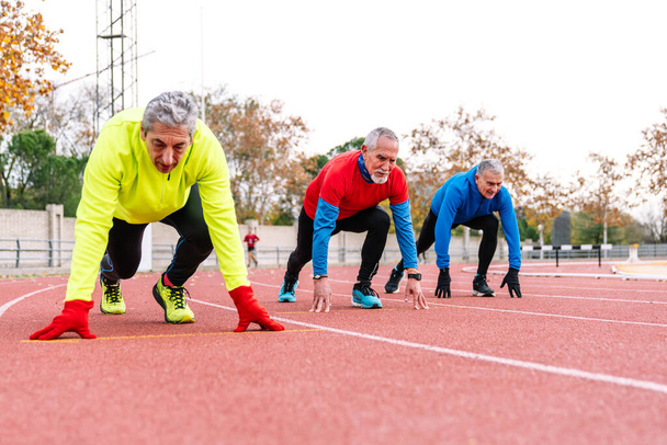 Três homens seniores posicionados e focados na linha de partida em uma pista, incorporando espírito competitivo e fitness em seus anos dourados. - Foto, Imagem