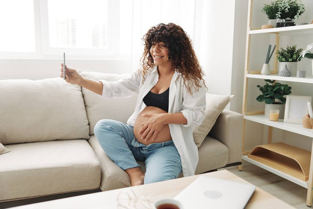 Raskaana oleva nainen bloggaaja istuu sohvalla kotona ja ottaa kuvia itsestään puhelimessa, selfiessä ja videopuhelussa, lääkärin kanssa verkossa, raskauden hallinnassa. Laadukas kuva - Valokuva, kuva