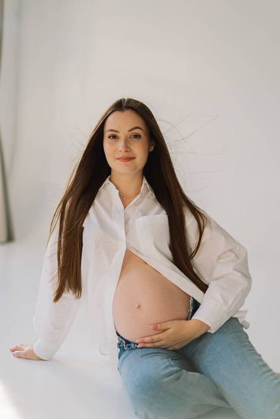 Cute młoda dziewczyna w ciąży siedzi na białym studio w ciepłym świetle z rąk na brzuchu w ciąży na tle ściany z cieniem. Koncepcja ciąży, wolna przestrzeń, cieszenie się ciążą. Ciało w ciąży. - Zdjęcie, obraz
