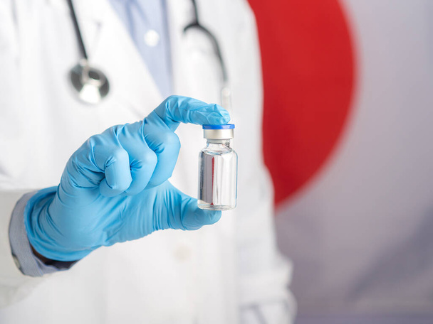 The doctor holding a vaccine bottle prepares vaccination against the background of the Japanese flag. Vaccine for immunization and treatment from virus infection. Medical concept. - Photo, Image