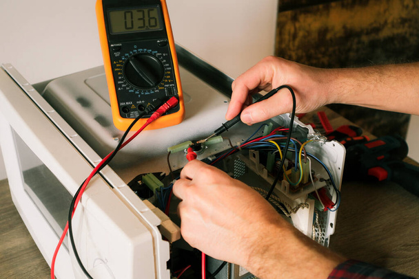 Un ingeniero de reparación utiliza un multímetro para comprobar los circuitos de un horno de microondas, en busca de una falla. Diagnósticos reparación de horno microondas. - Foto, imagen