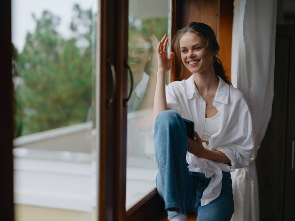 Jeune femme assise sur un rebord de fenêtre en bois près de la fenêtre avec un téléphone à la main souriant et regardant par la fenêtre, humeur d'automne, atmosphère confortable à la maison, style de vie esthétique. Photo de haute qualité - Photo, image