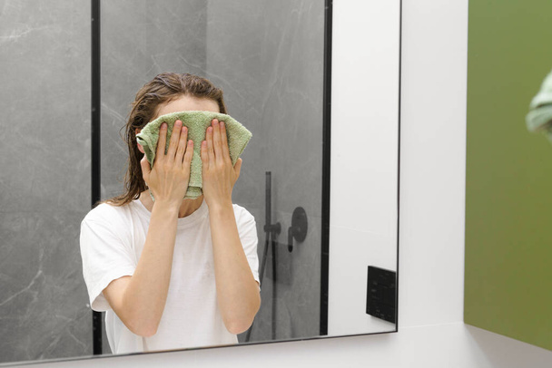 Молодая женщина вытирает лицо зеленым полотенцем после мытья в ванной. Концепция ежедневной гигиены, ухода за лицом и кожей - Фото, изображение