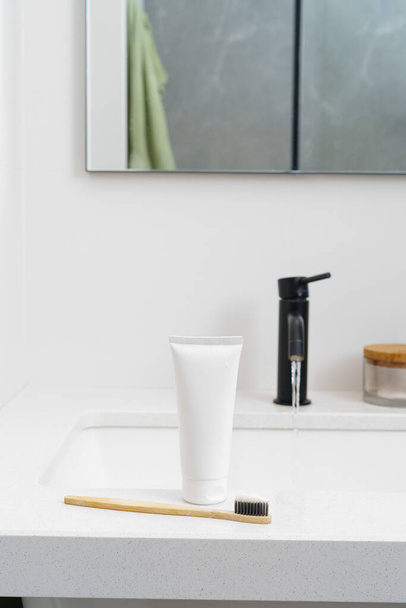 歯磨き粉のチューブと木製の歯ブラシの白いモックアップは,タップからの水の流れの背景のシンクの上に立っています. 縦のイメージ。 自然衛生製品の概念,口腔のケア. - 写真・画像