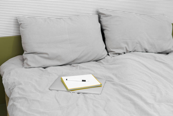 Un portátil plegado gris y un cuaderno con una pluma se encuentran en la ropa de cama. Concepto de trabajo y aprendizaje a distancia, búsqueda de información en red - Foto, imagen